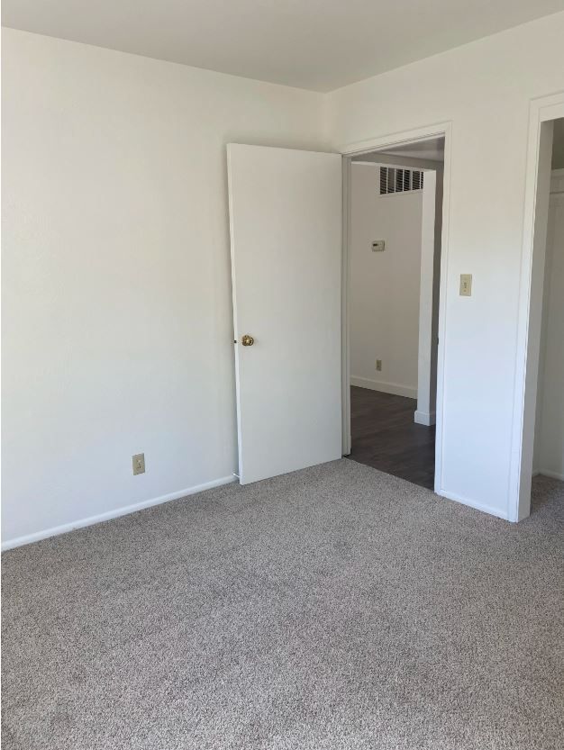 Empty room with white walls, closed door, and neutral carpet. Another doorway leads to a hallway.