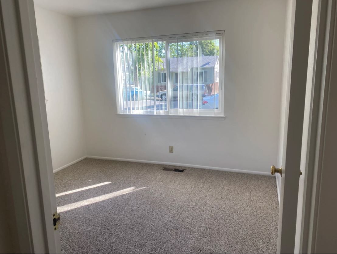 Empty room with a window, white walls, and carpet, viewed from a doorway. Sunlight streams through the window.