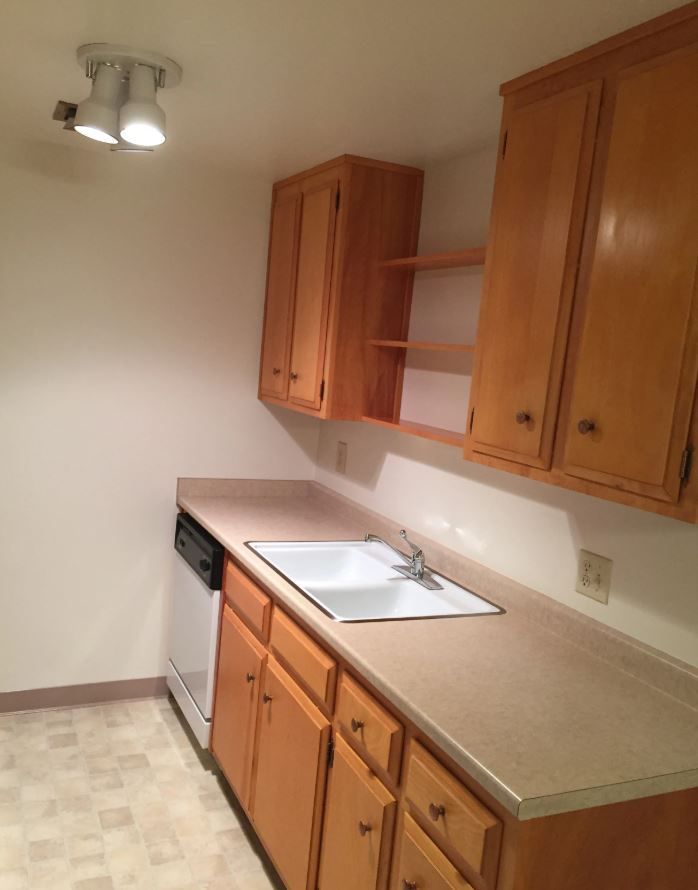 A small kitchen with wood cabinets, a white sink, and a dishwasher. Beige countertops and flooring.