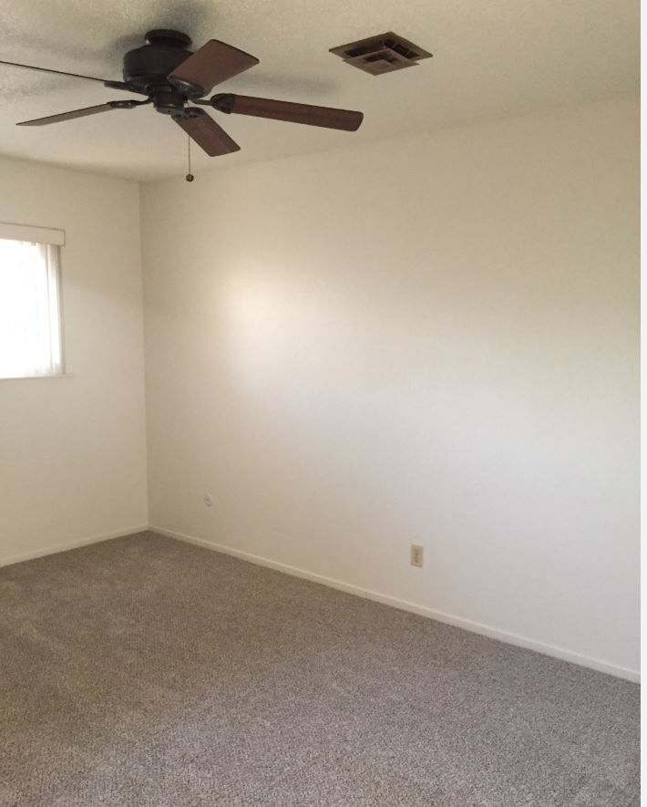 Empty bedroom with tan carpet, white walls, ceiling fan, small window, and vent.
