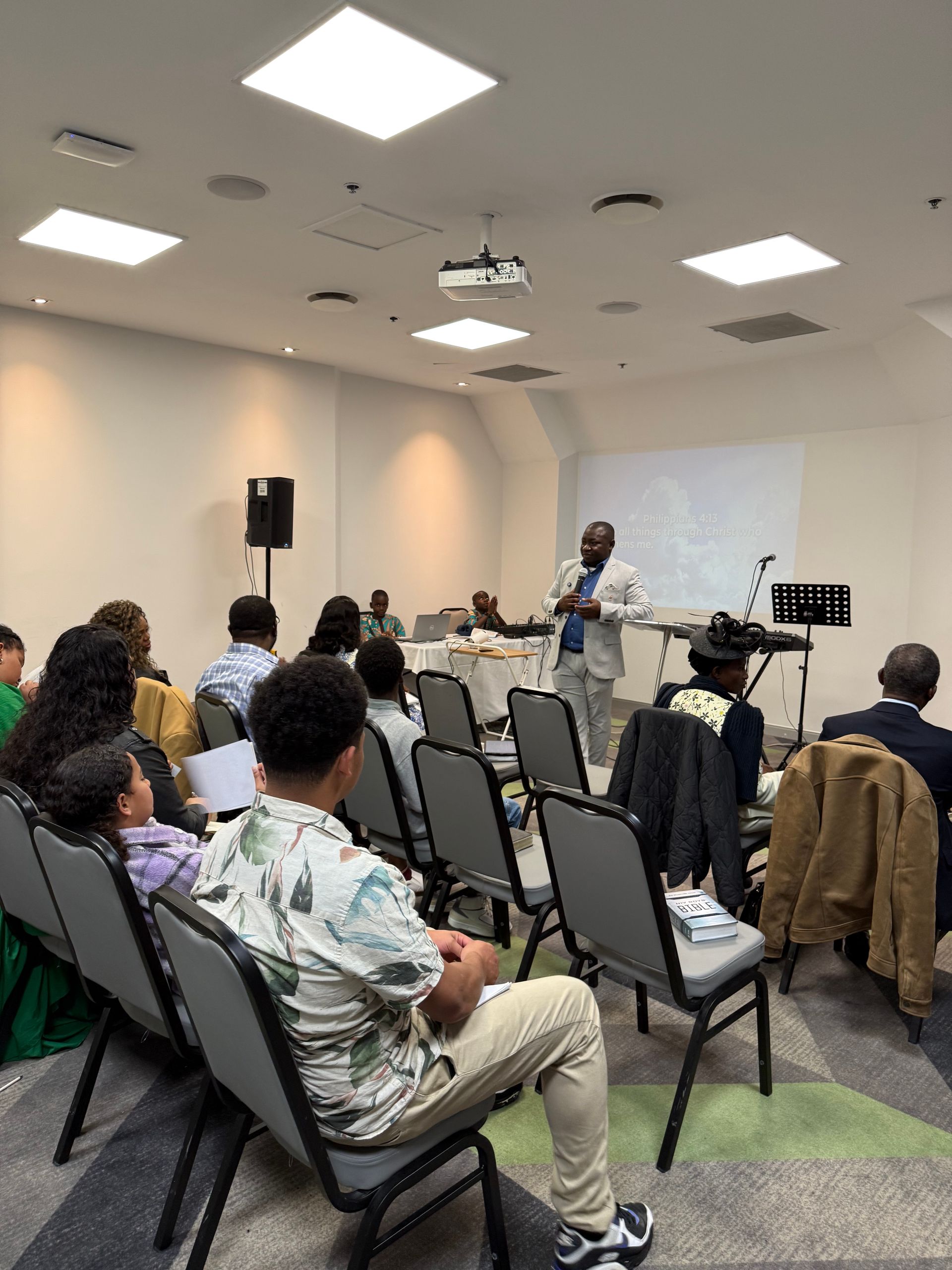 A man speaks at a church service. Attendees sit in chairs facing him. White walls and bright lights.