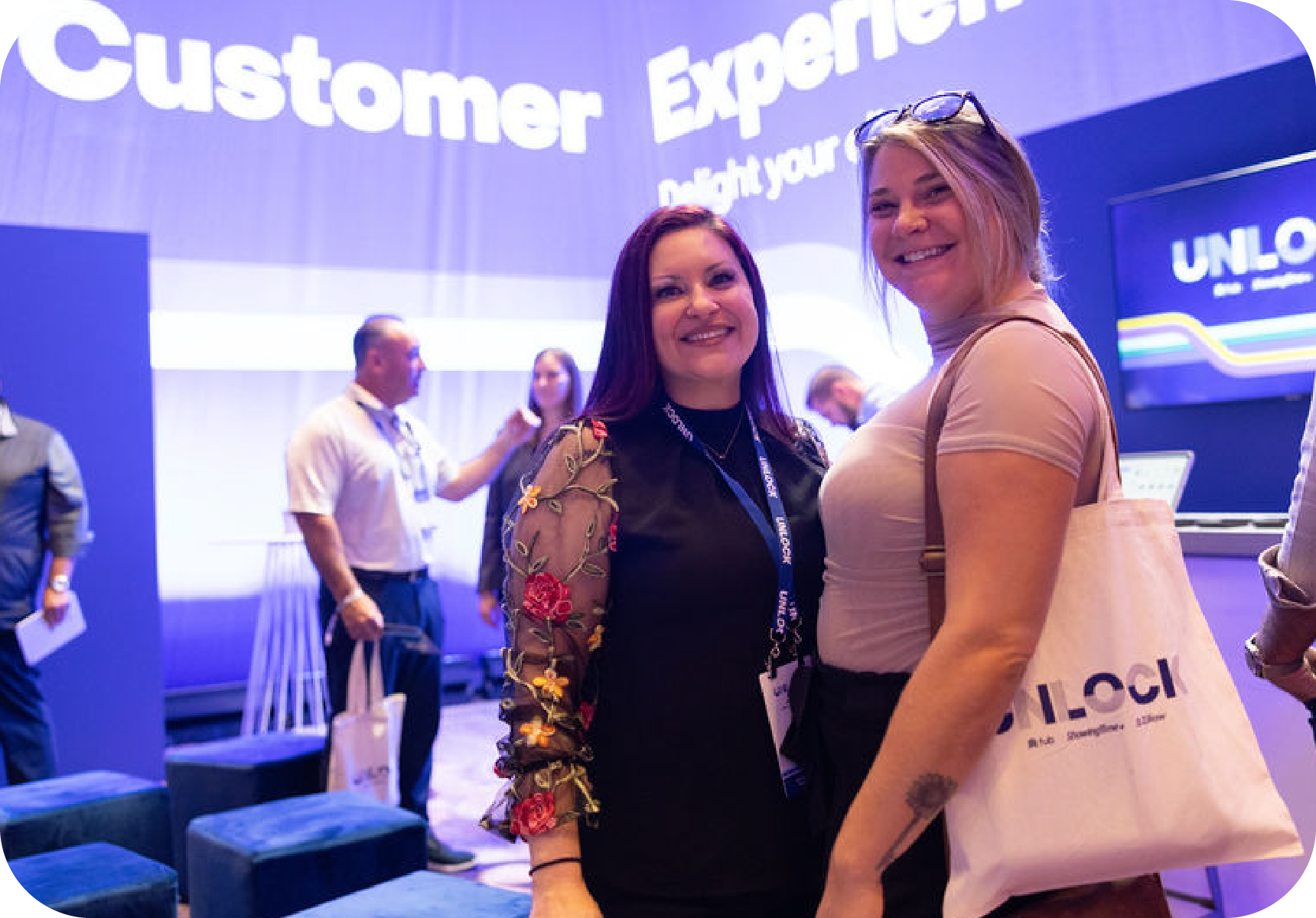 Two women smiling at a trade show booth. One has purple hair, the other holds a tote bag. People and a screen are in the background.