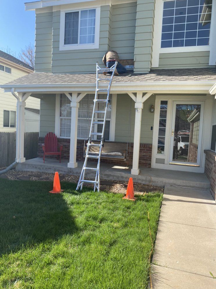 A person on a ladder repairs the exterior wall of a light green, two-story house with orange safety cones on the lawn.