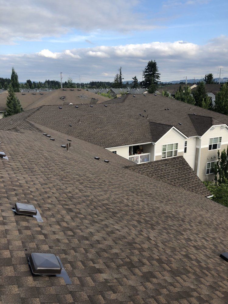 View from a rooftop overlooking a multi-story apartment complex with brown asphalt shingles under a partly cloudy sky.