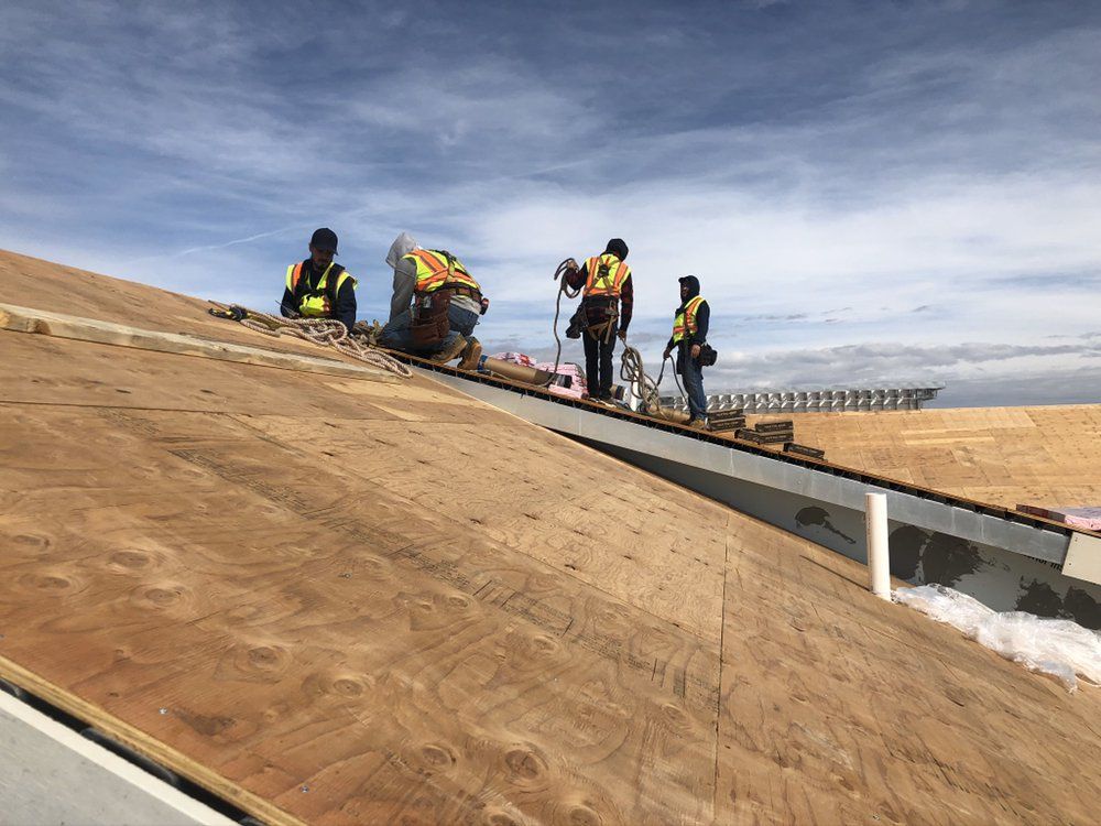 Four construction workers in safety vests install shingles on a sloped residential roof under a cloudy sky.