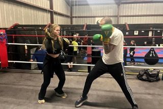 A woman and a man boxing in a gym. The woman throws a punch, while the man, wearing green gloves, is in a defensive stance.
