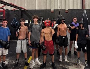 Group of young boxers in a gym, some shirtless, wearing gloves and protective headgear, posing for the camera.