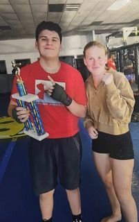 A young man in a red shirt holding a trophy, and a young woman in a tan jacket and black shorts pose in a gym.