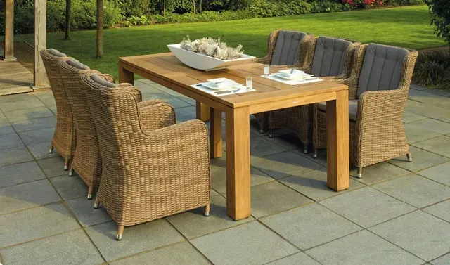 Light brown patio table with six wicker chairs in a backyard sitting on a large grey square paver patio.