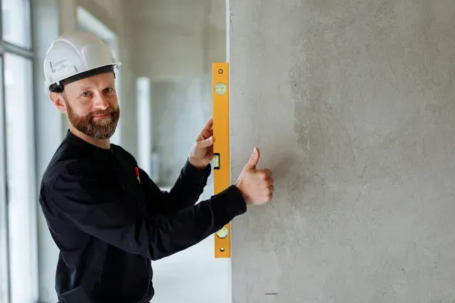 Contractor wearing a long-sleeve black shirt and a white hard hat holding a 2-foot yellow level vertically against a wall in a house.