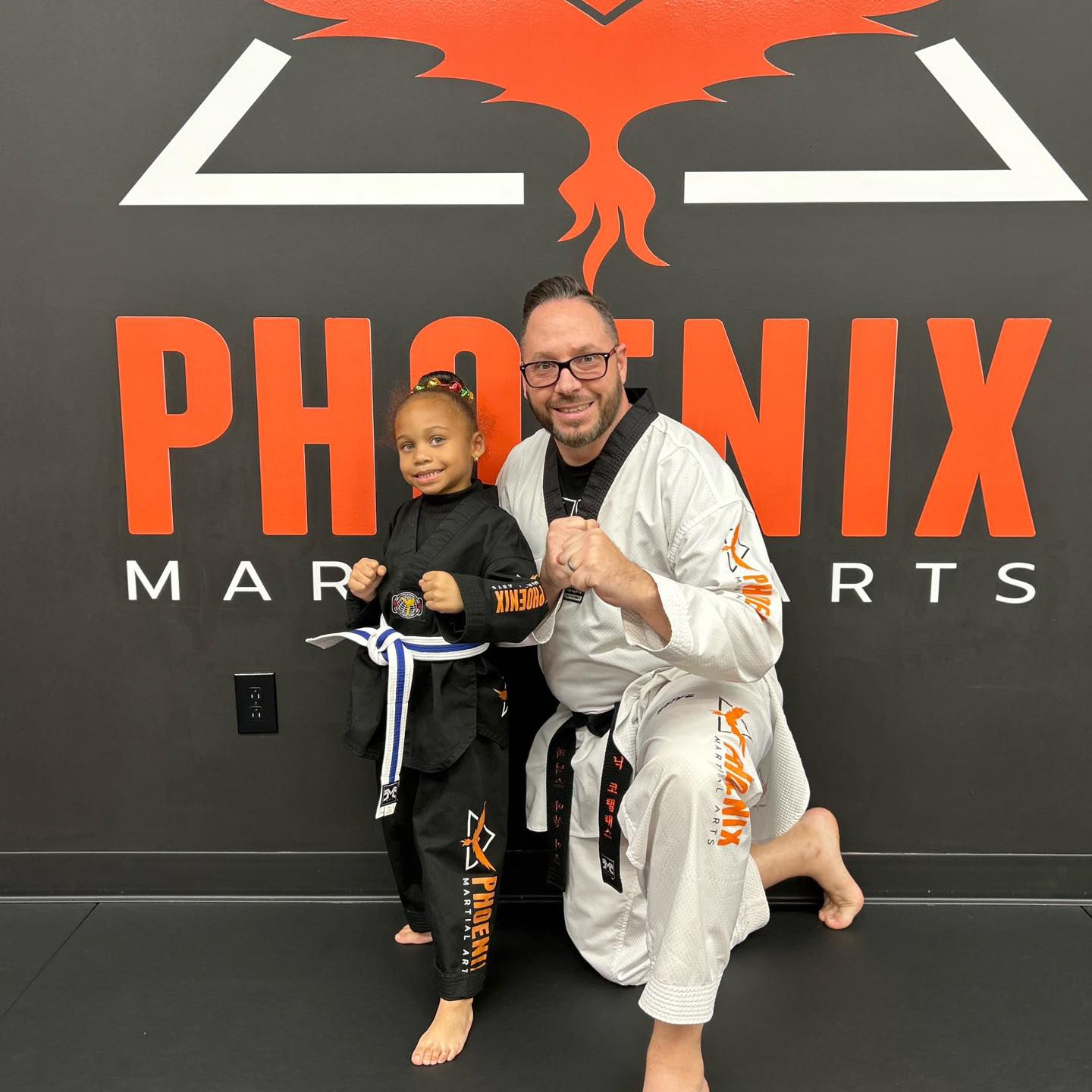 A man and a little girl are posing for a picture in front of a phoenix martial arts sign.