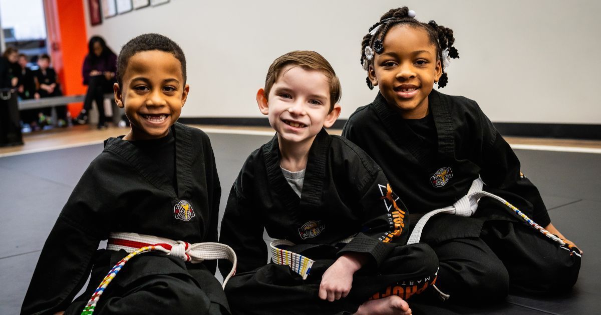 Three young children are sitting on the floor in karate uniforms.