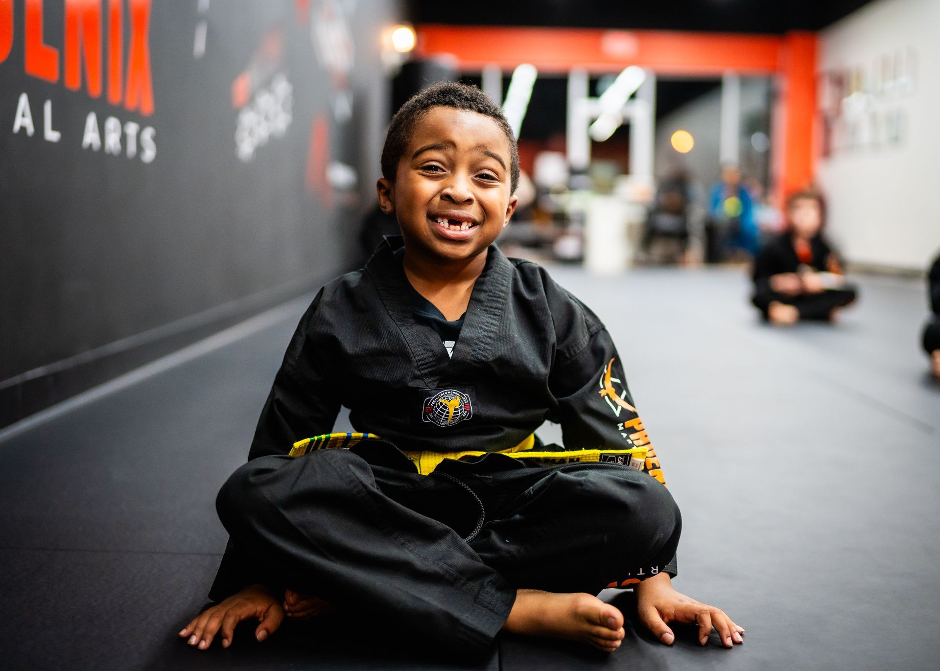 A young boy in a karate uniform is sitting on the floor in a gym.