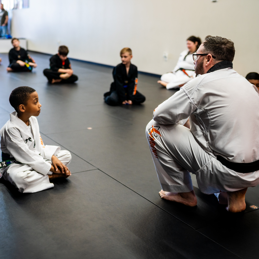 A man is kneeling down next to a young boy in a taekwondo class.