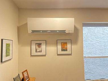 Air conditioning unit mounted on a beige wall, with framed artwork and a window.