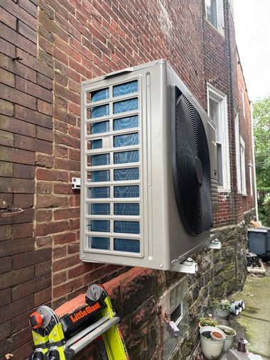 Outdoor HVAC unit mounted on a brick wall, with a ladder and potted plants visible below.