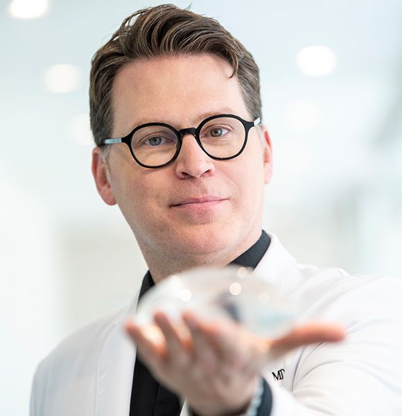 Un homme portant des lunettes et une blouse blanche tient un verre à la main.