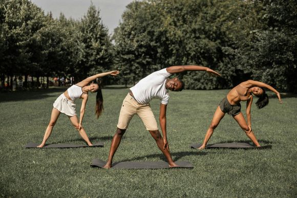 Three people in athletic wear perform a side-stretch yoga pose on mats in a grassy park surrounded by trees.