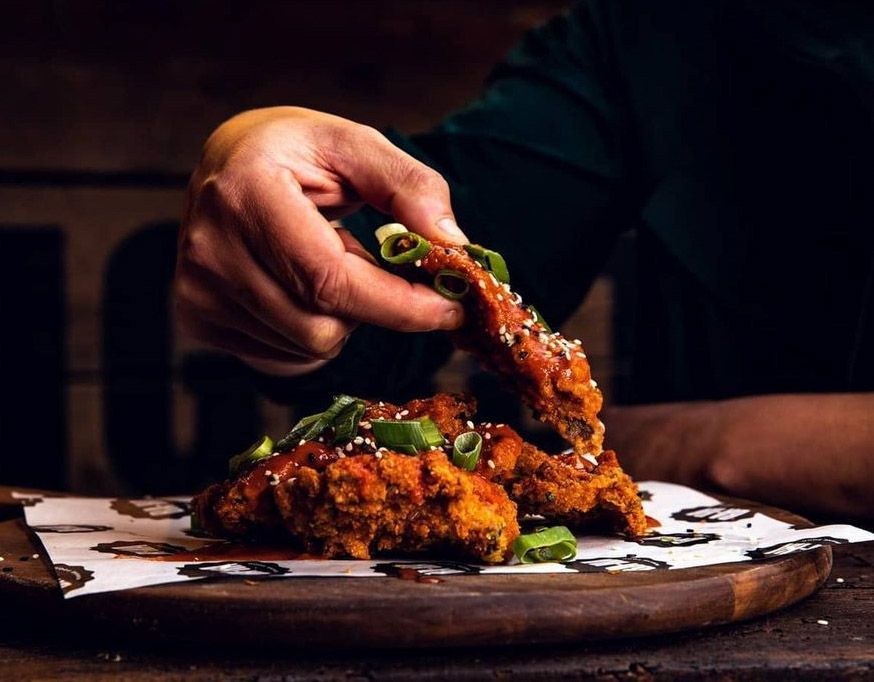Hand holding a piece of fried chicken topped with green onions and sesame seeds. Chicken on a wooden board.