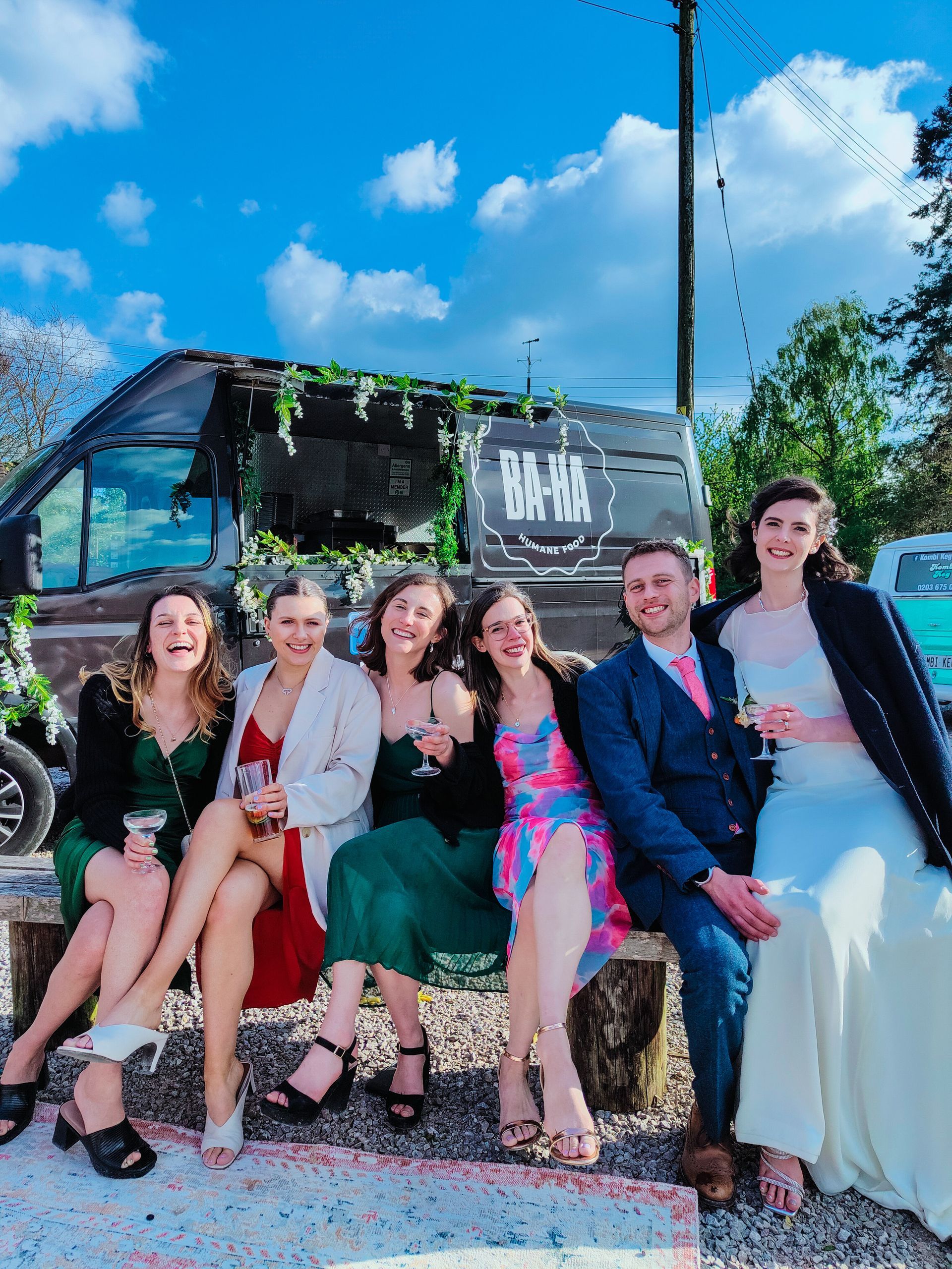 Six people, in formal wear, sit on a bench in front of a van, smiling and holding drinks.