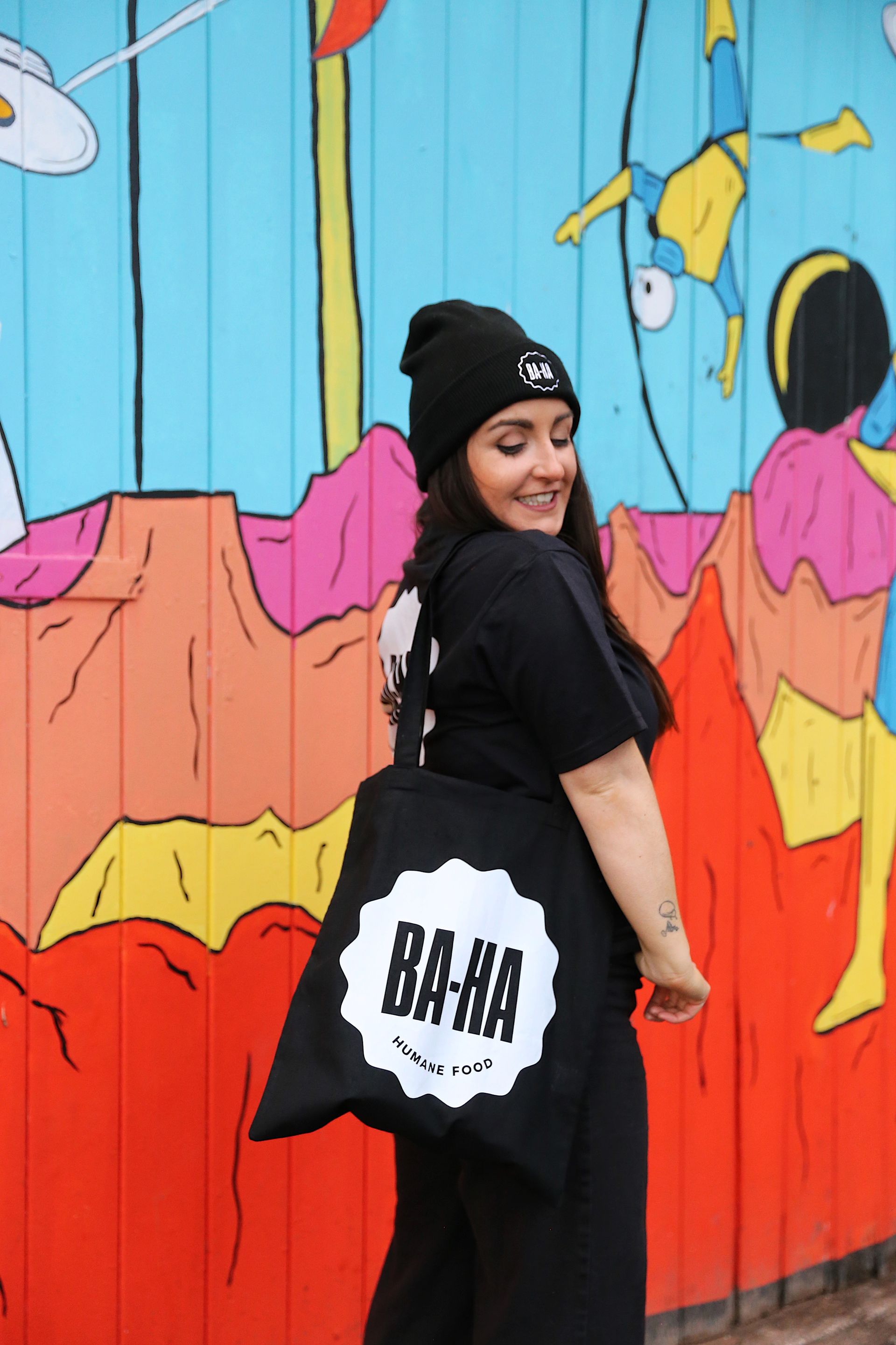 Woman wearing black beanie, t-shirt, and carrying a tote bag, smiles in front of a colorful mural.