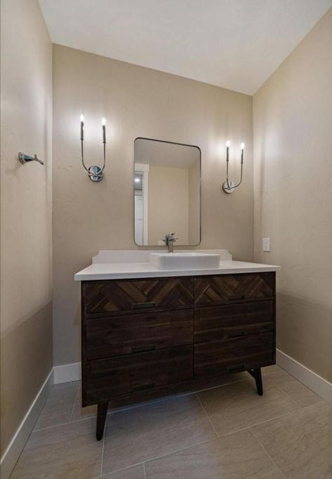 Small bathroom vanity with dark wood cabinet, white sink, mirror, and wall sconces on beige walls.