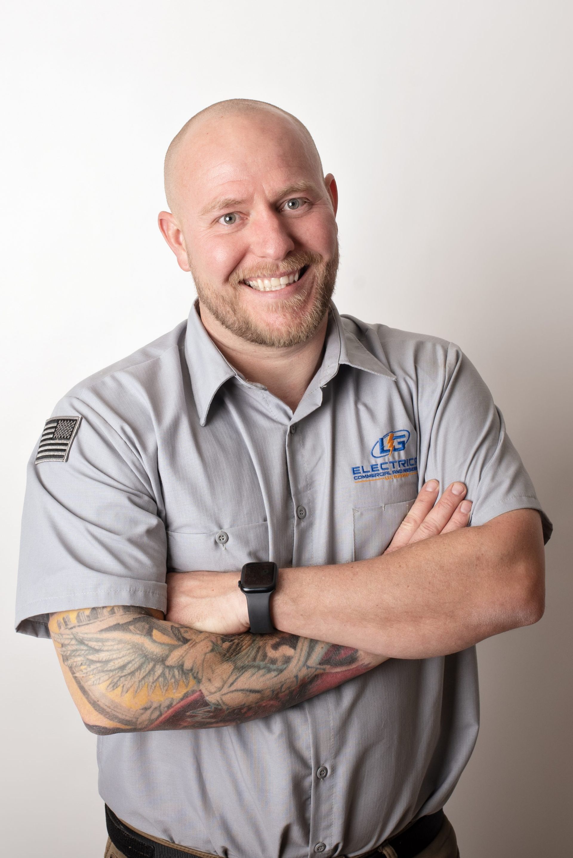 Bald man with crossed arms, tattoos, and a gray work shirt smiles at the camera.