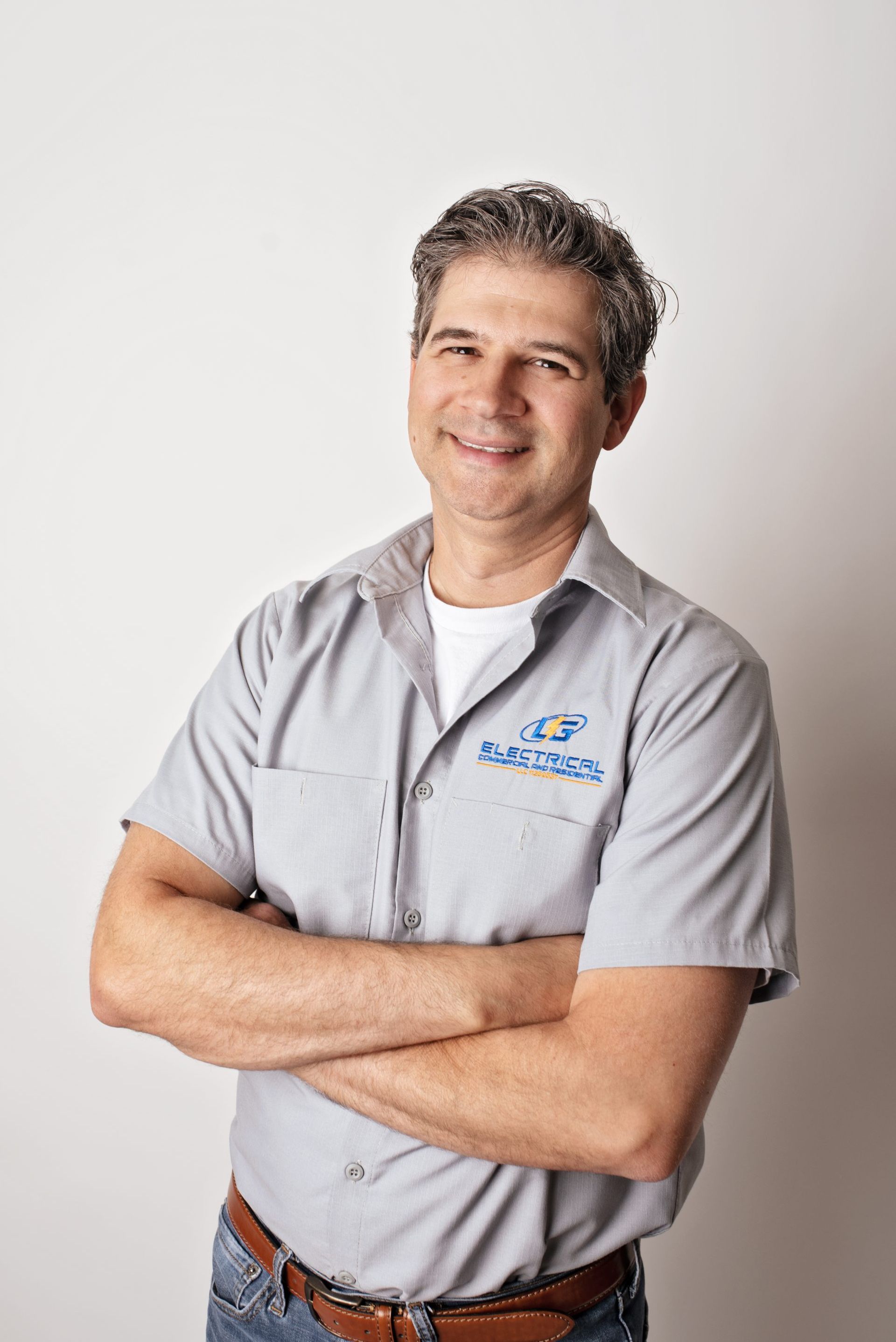 Man with arms crossed, wearing a grey work shirt and jeans, smiling in front of a white background.