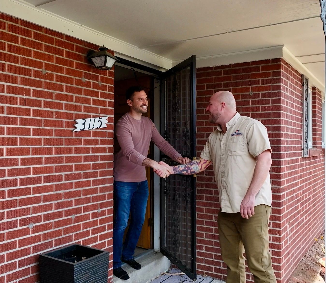 Two men shaking hands at a brick house doorway. One is inside the door, smiling. The other is on the porch.