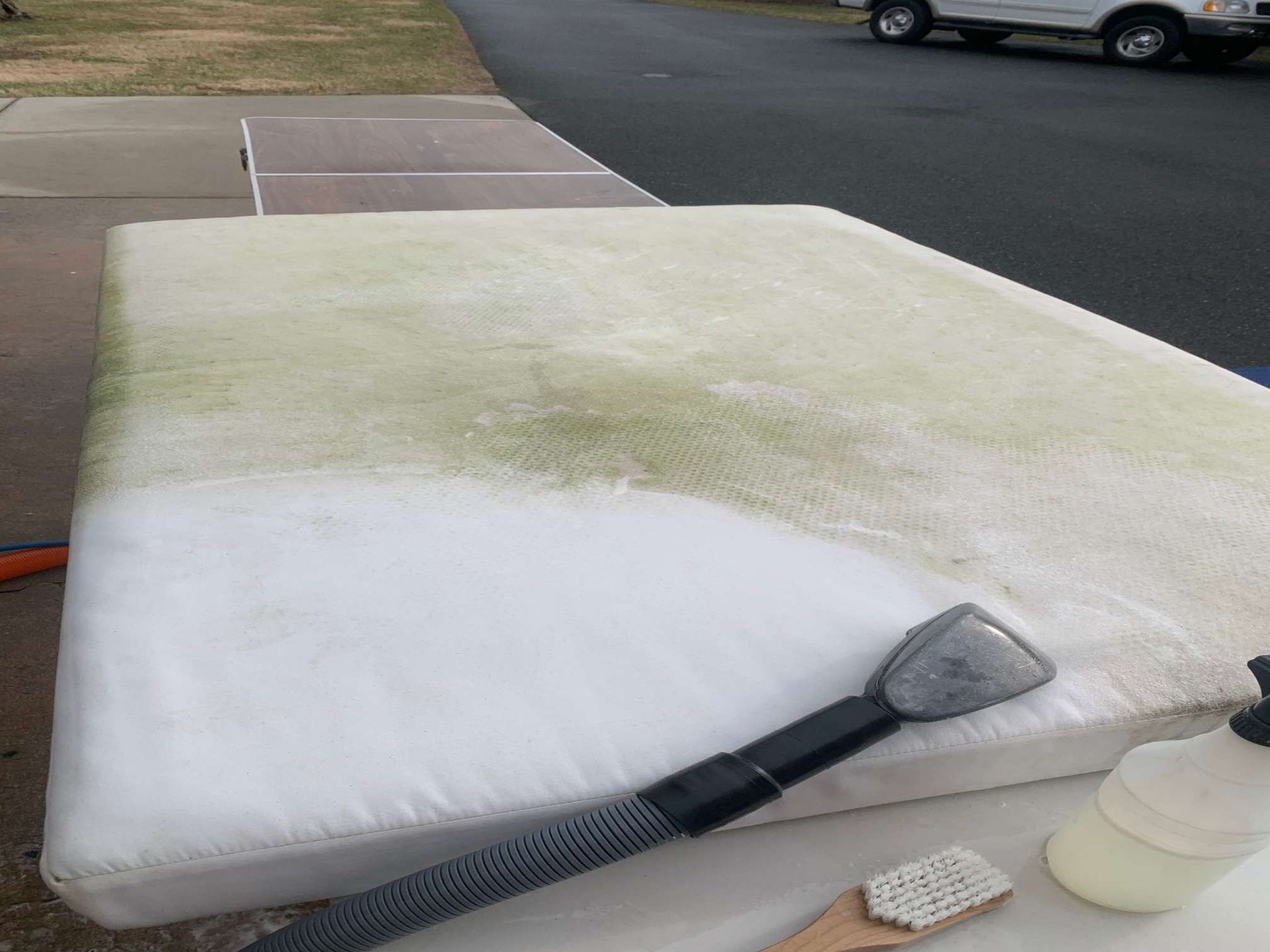 A partially cleaned, stained mattress outdoors, with a vacuum extraction tool, scrub brush, and cleaning spray nearby.