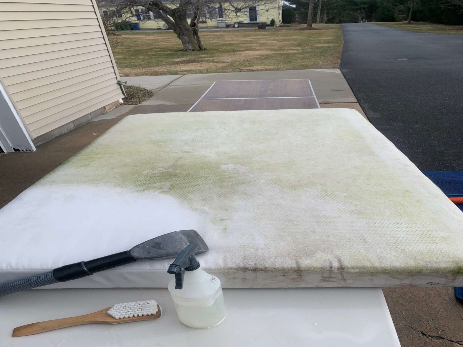 A dirty outdoor cushion is partially cleaned with a white cleaning solution, a scrub brush, and a squeegee on a table.