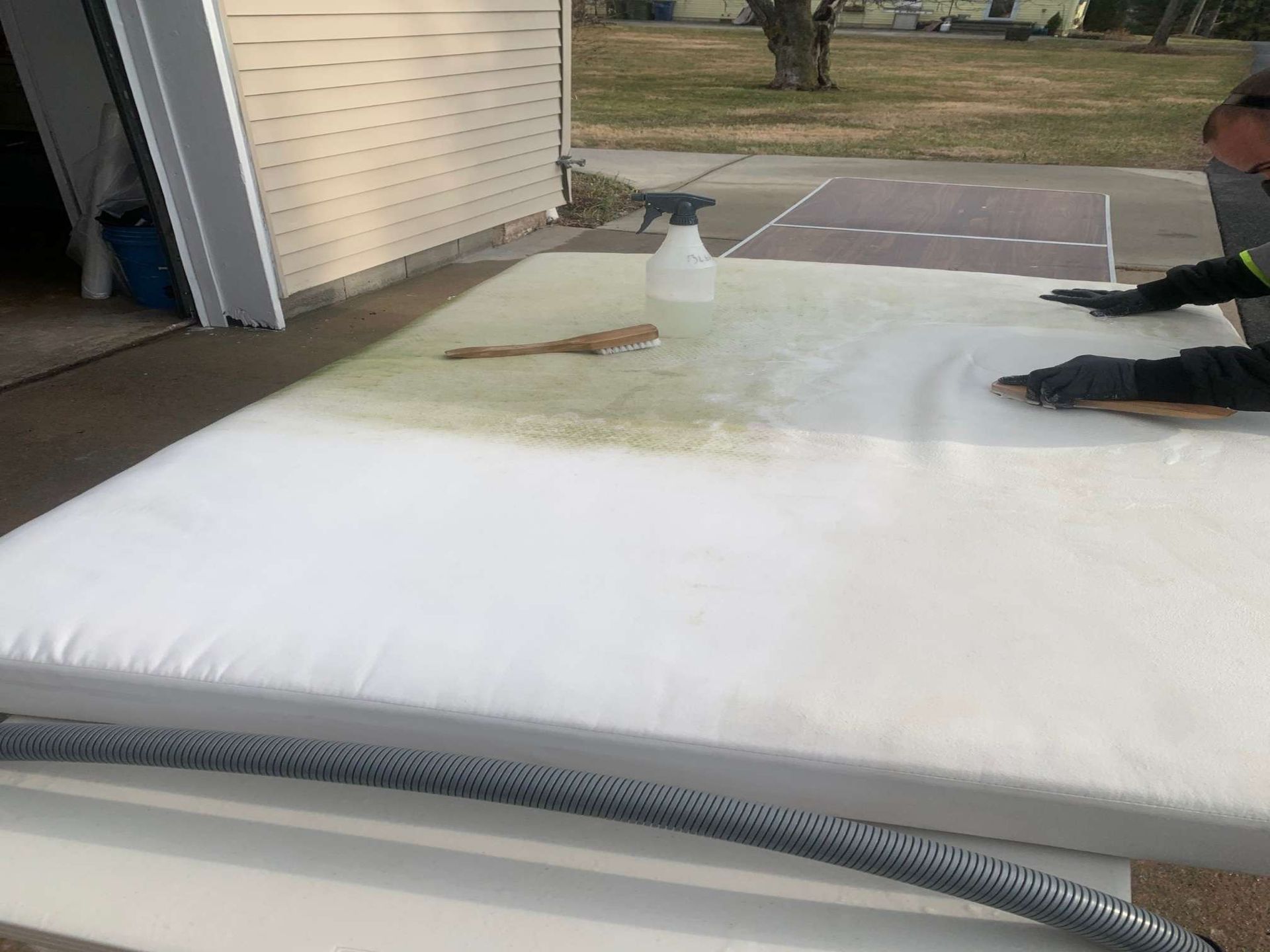 A person wearing gloves scrubs a large white mattress outdoors with a cleaning brush and spray bottle.
