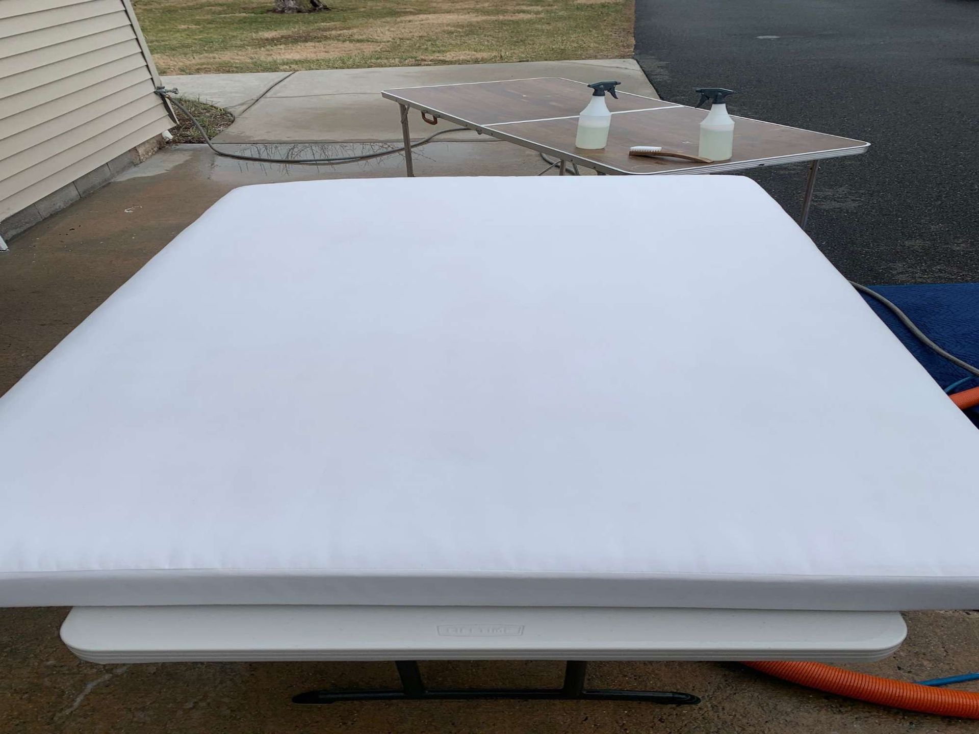 A white, padded outdoor cushion rests on a table, with cleaning supplies visible on a separate table in the background.