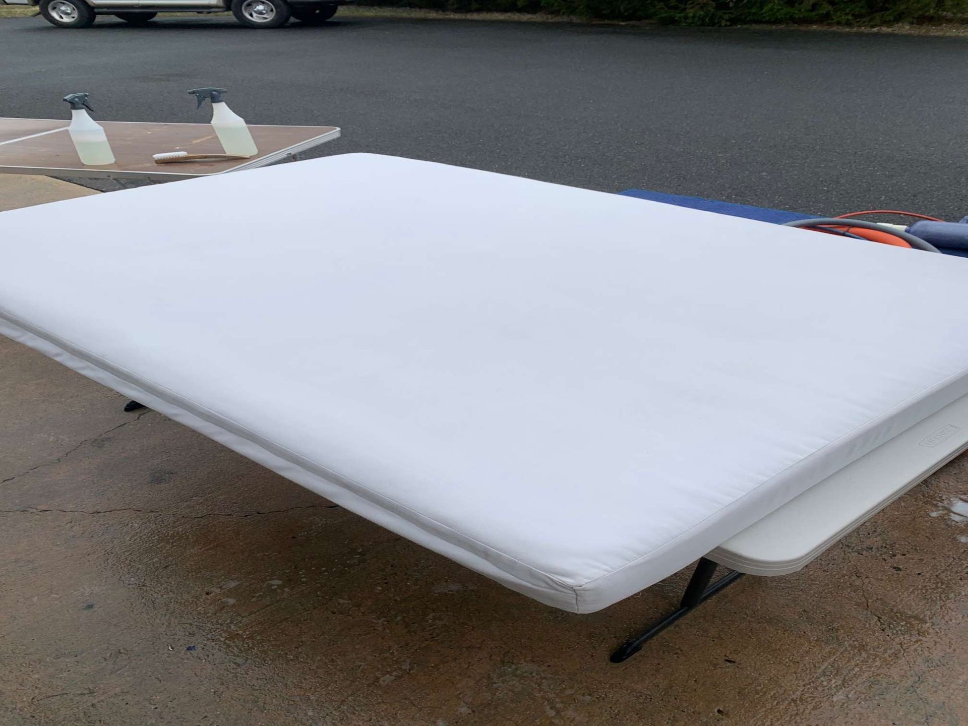 A white mattress sits on a folding table on an asphalt surface, with two spray bottles nearby.