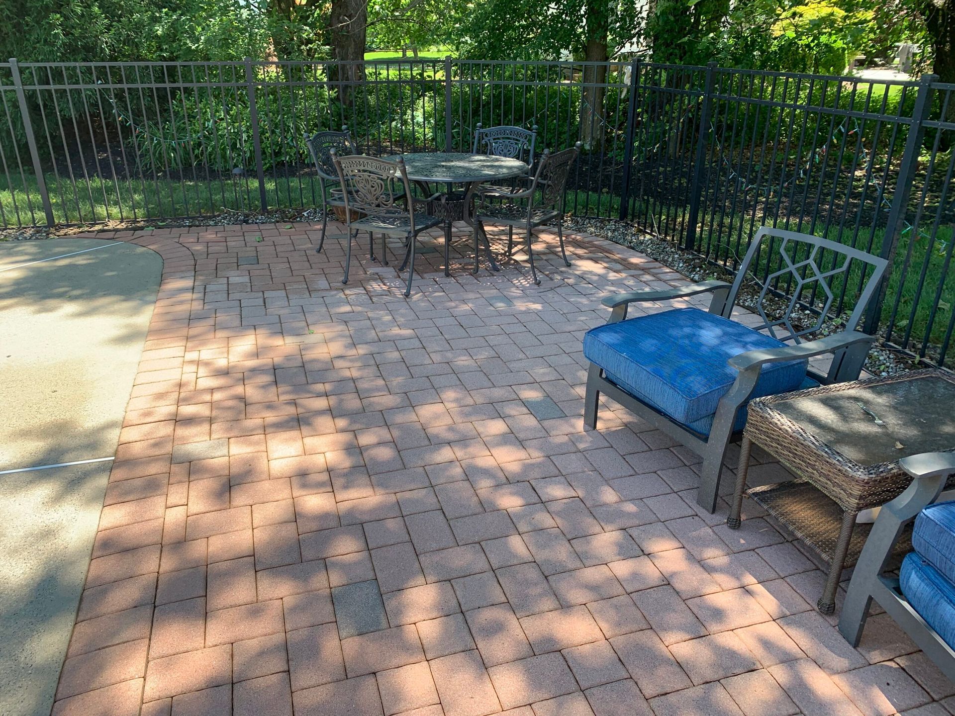 A patio with brick pavers, a wrought iron dining set, and comfortable armchairs with blue cushions by a metal fence.