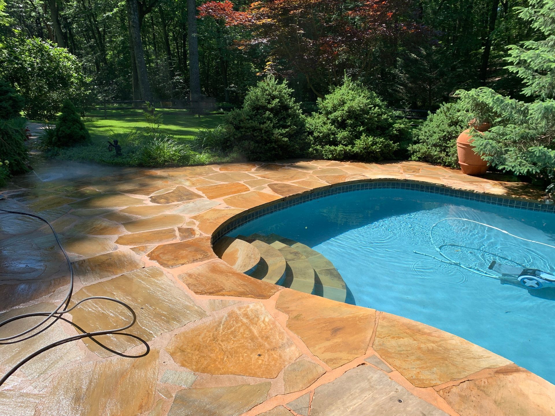 A flagstone pool deck with steps leading into a blue swimming pool, surrounded by green trees and shrubs.