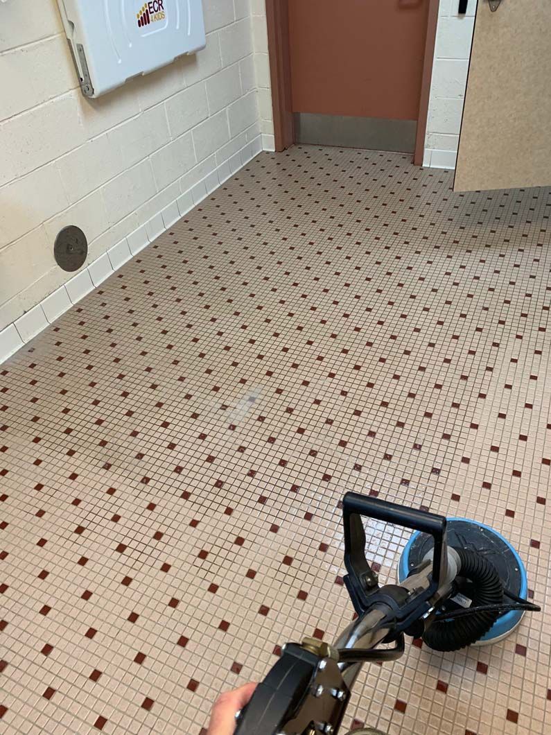 A person using a floor buffer machine on a light-colored tiled floor with a repeating small dark square pattern.
