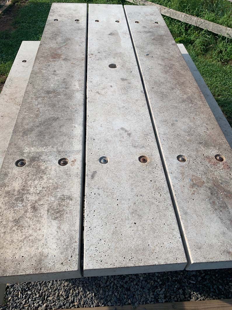 A top-down view of a light gray concrete picnic table surface, showing three rectangular planks with bolt-head fasteners.