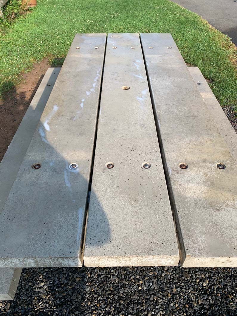 A rectangular, light gray, three-plank concrete picnic table sits outdoors on gravel near green grass.
