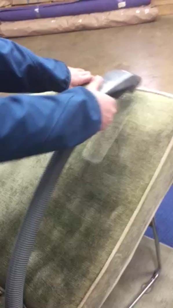 A person in a blue jacket uses an upholstery cleaning machine to clean a dirty, olive-green fabric surface.