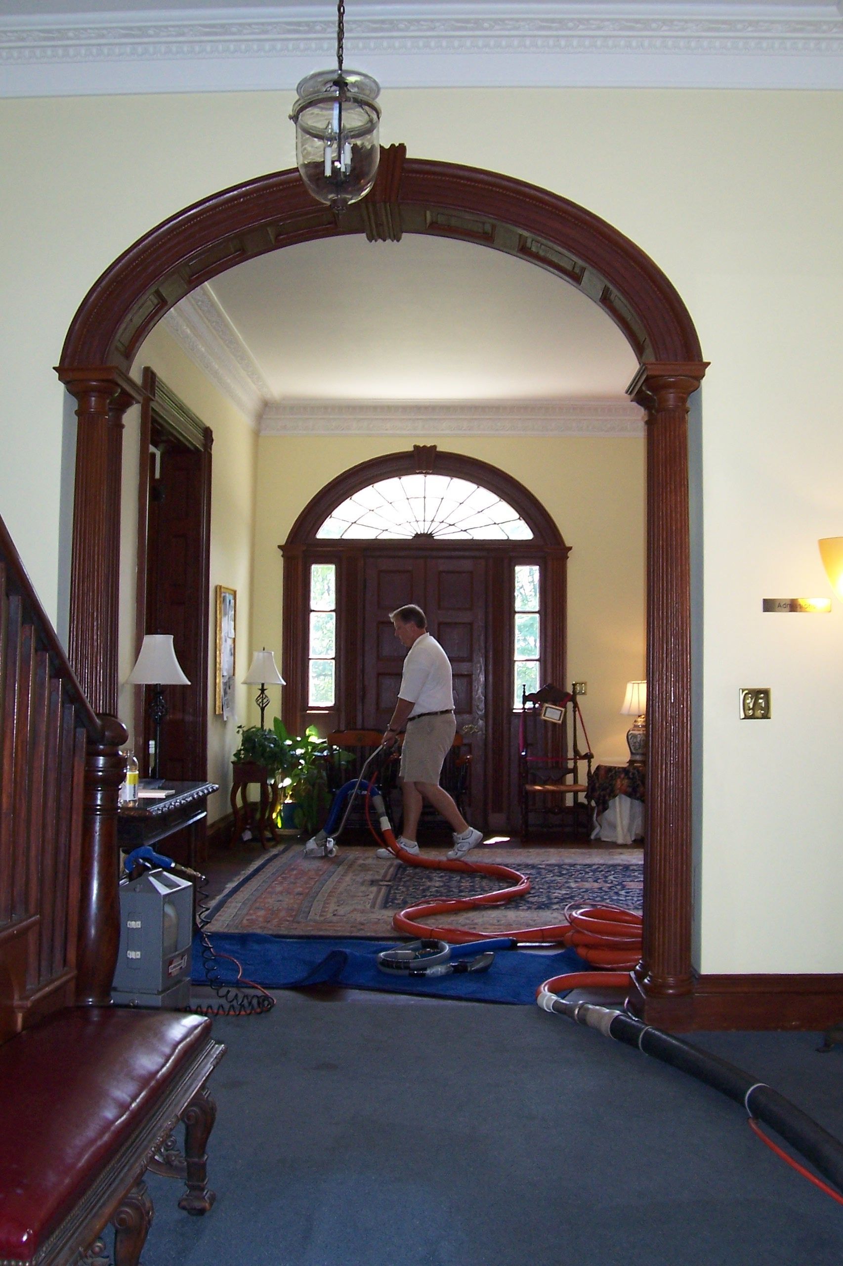 A worker cleans a rug in a grand entryway with an arched wooden doorway and a view of the front door.