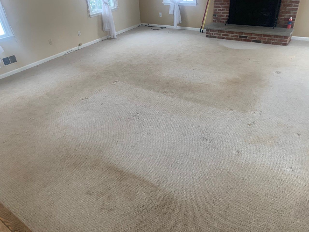 A light-colored carpet in a room with a brick fireplace and white curtains, showing significant uneven wear and dirt.