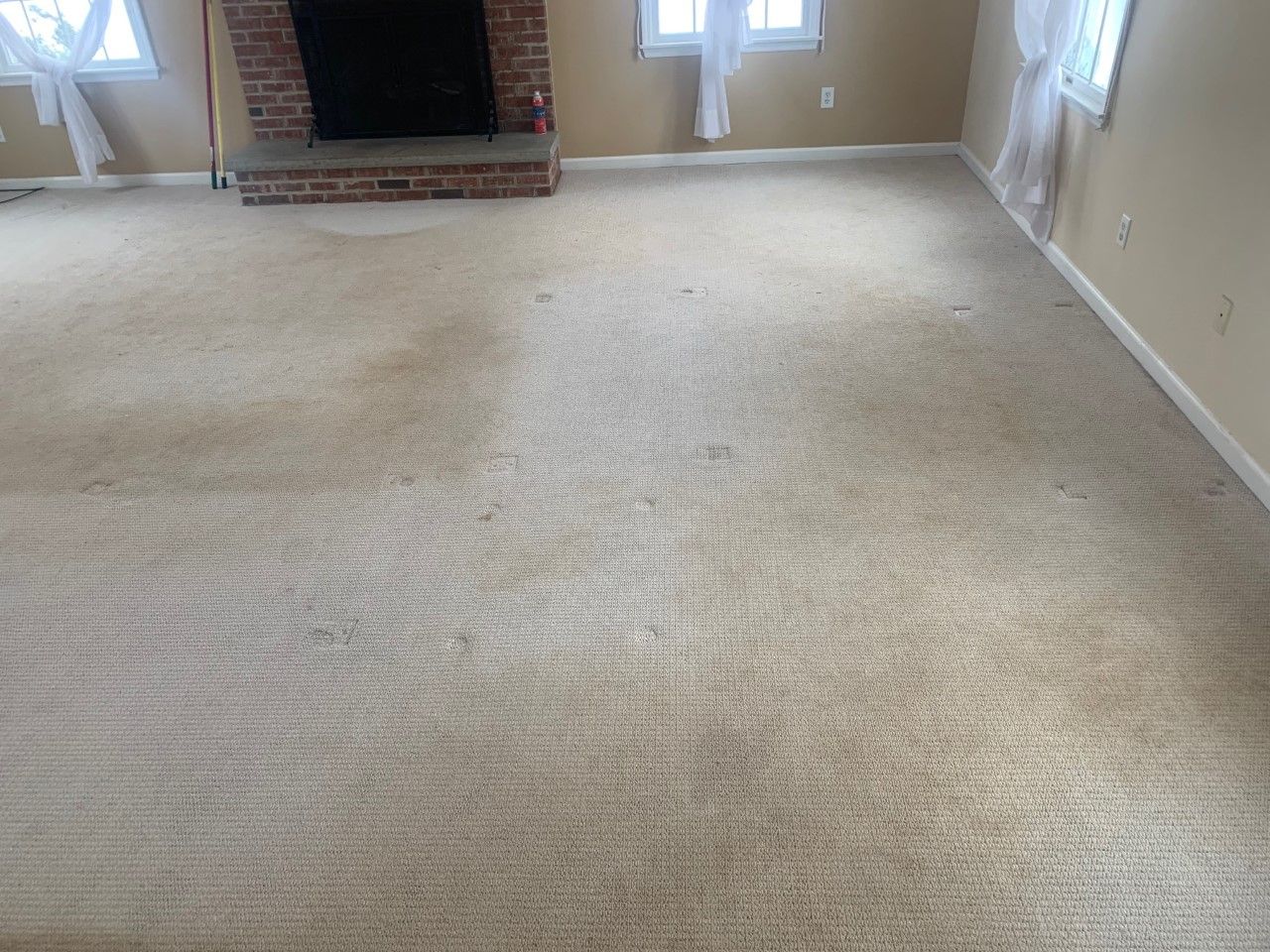 A beige, stained carpet covers a living room floor in front of a brick fireplace and several windows.