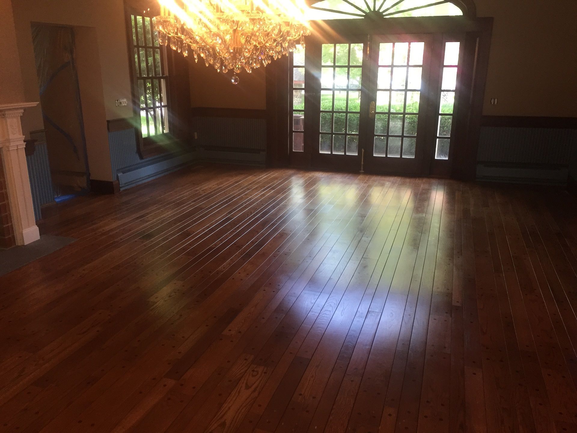 A sunlit room with dark wood flooring, a crystal chandelier, and large glass doors leading outdoors.