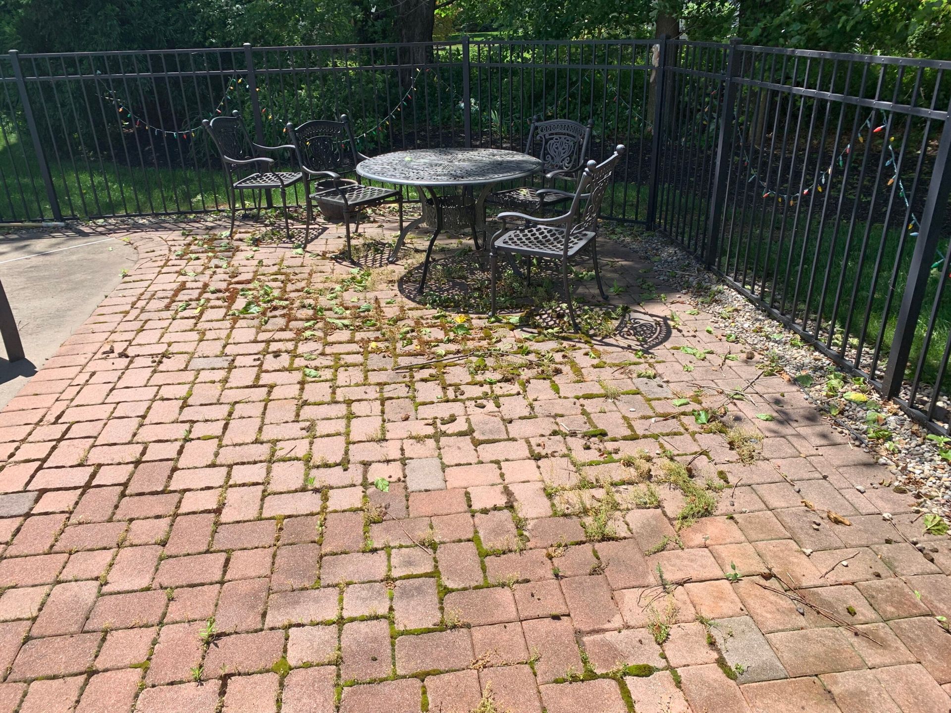 A patio with a round metal table and four matching chairs, surrounded by a black fence, with weeds growing in the pavers.