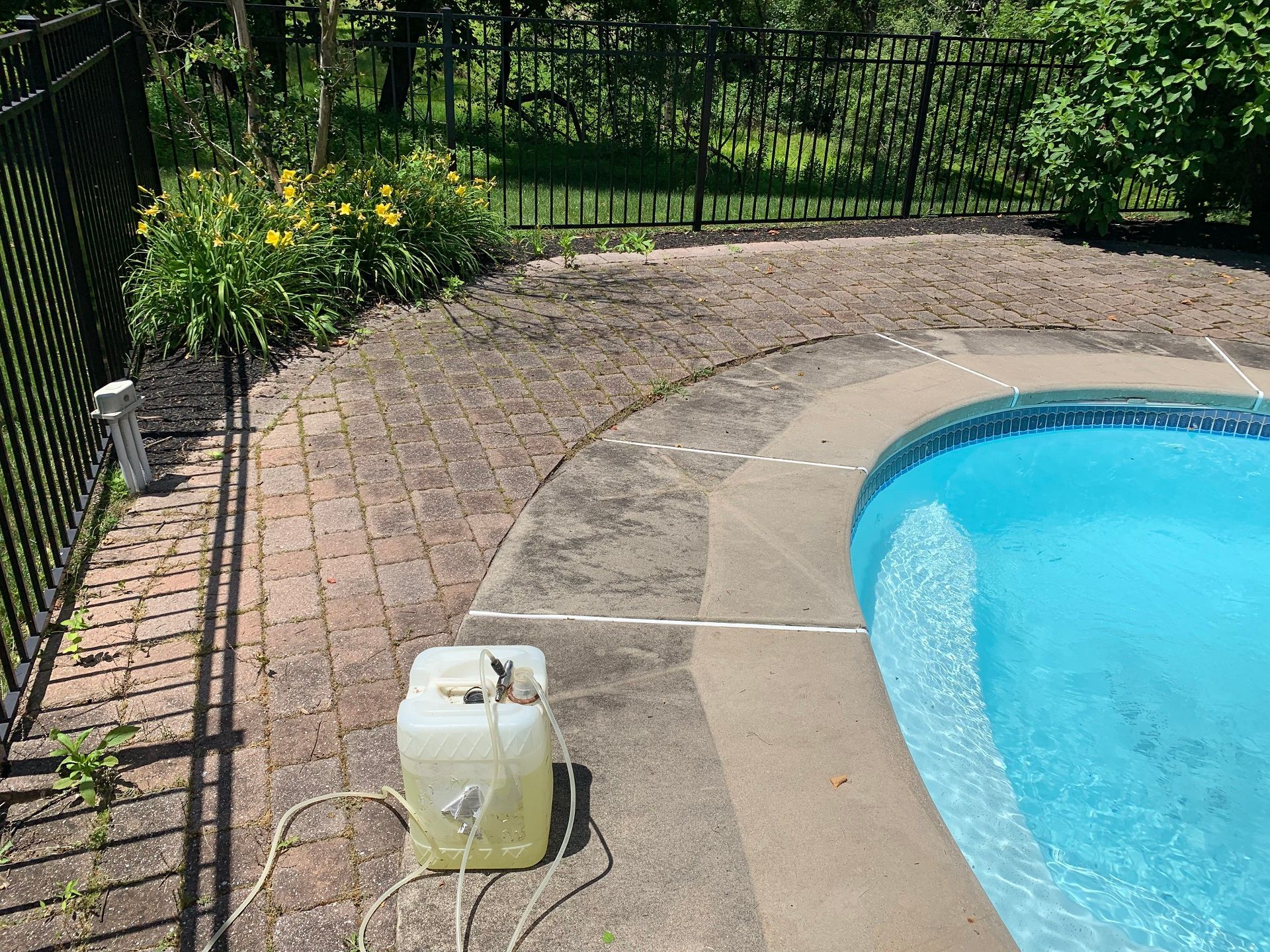 A plastic chemical container with hoses sits on a brick patio next to a swimming pool and a black metal fence.