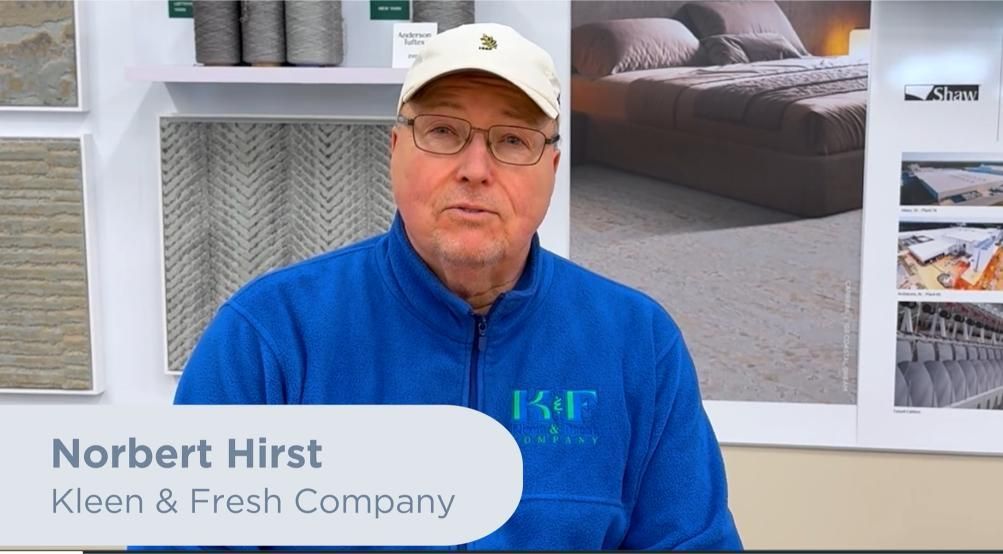 A person wearing a blue fleece with a logo and a cap stands in front of a display of carpets and home decor.