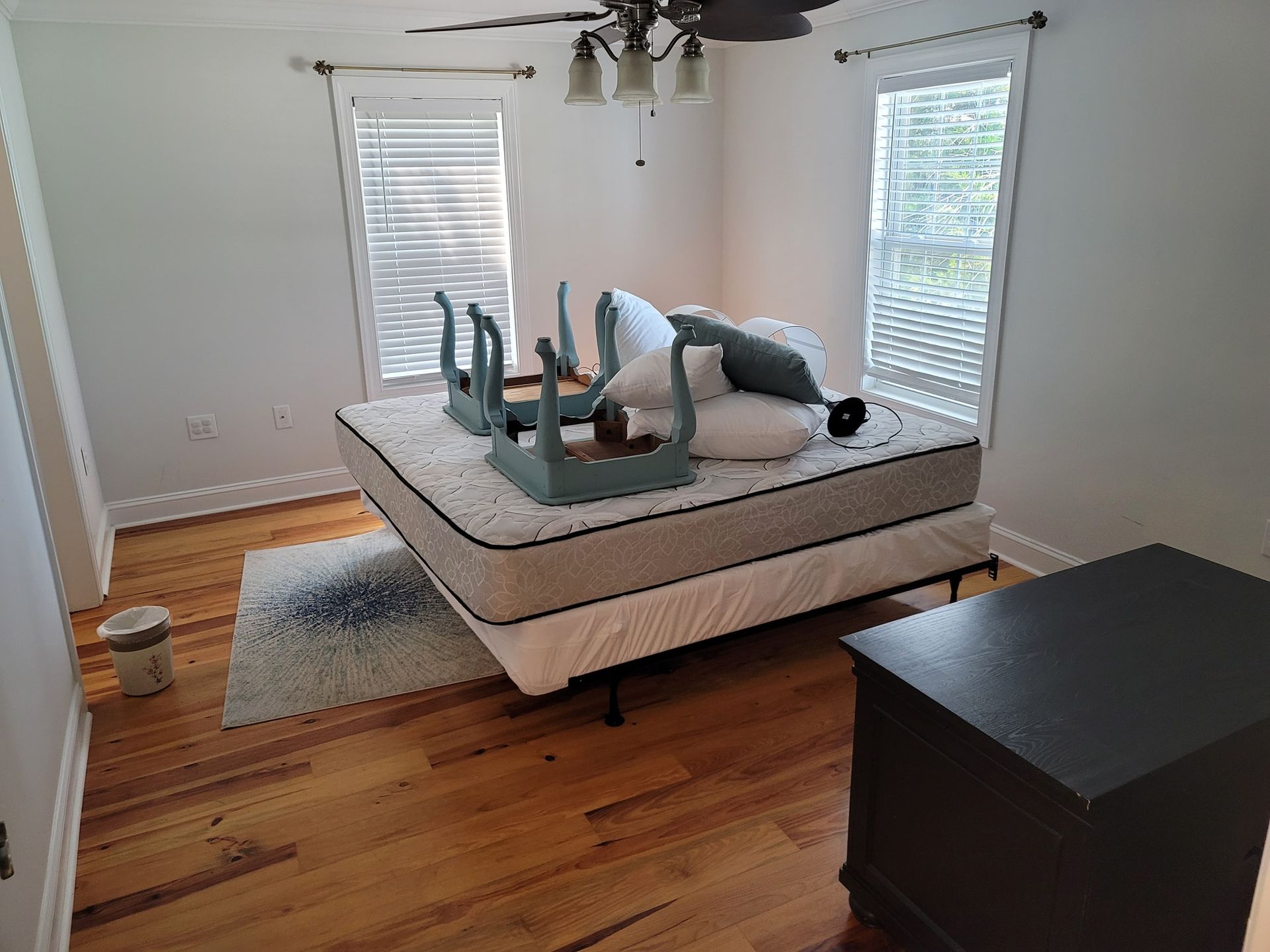 Bedroom with a bed, nightstand, chairs, and a rug. Furniture is placed on a hardwood floor, with windows and blinds.