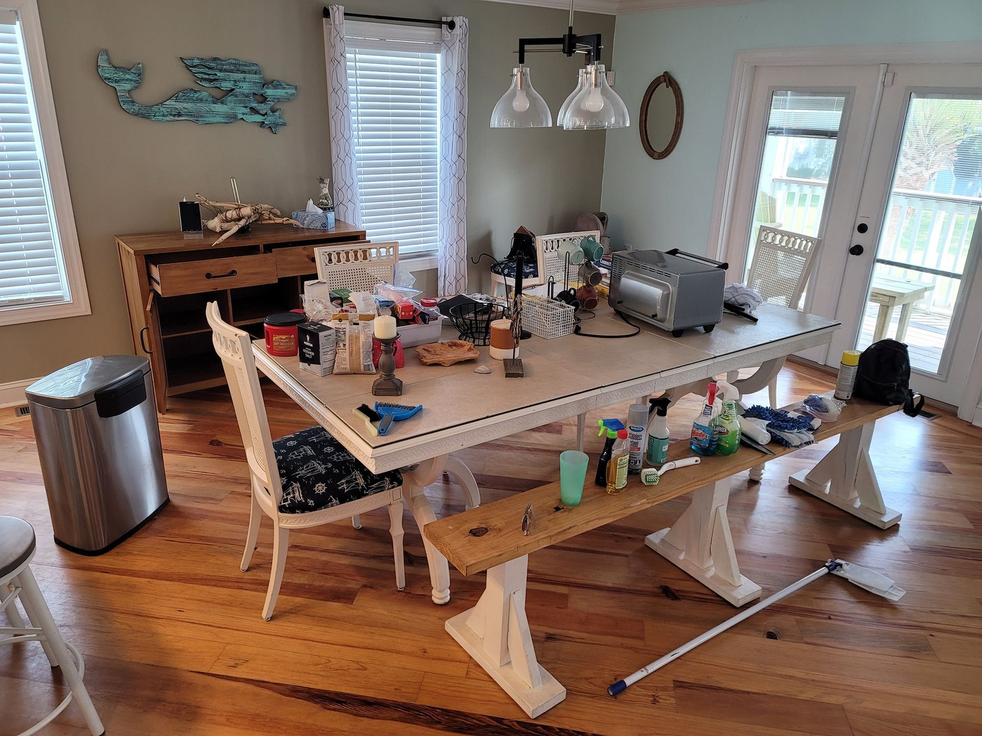 A cluttered dining room with a white table, wood floor, windows, and a stainless steel trash can.