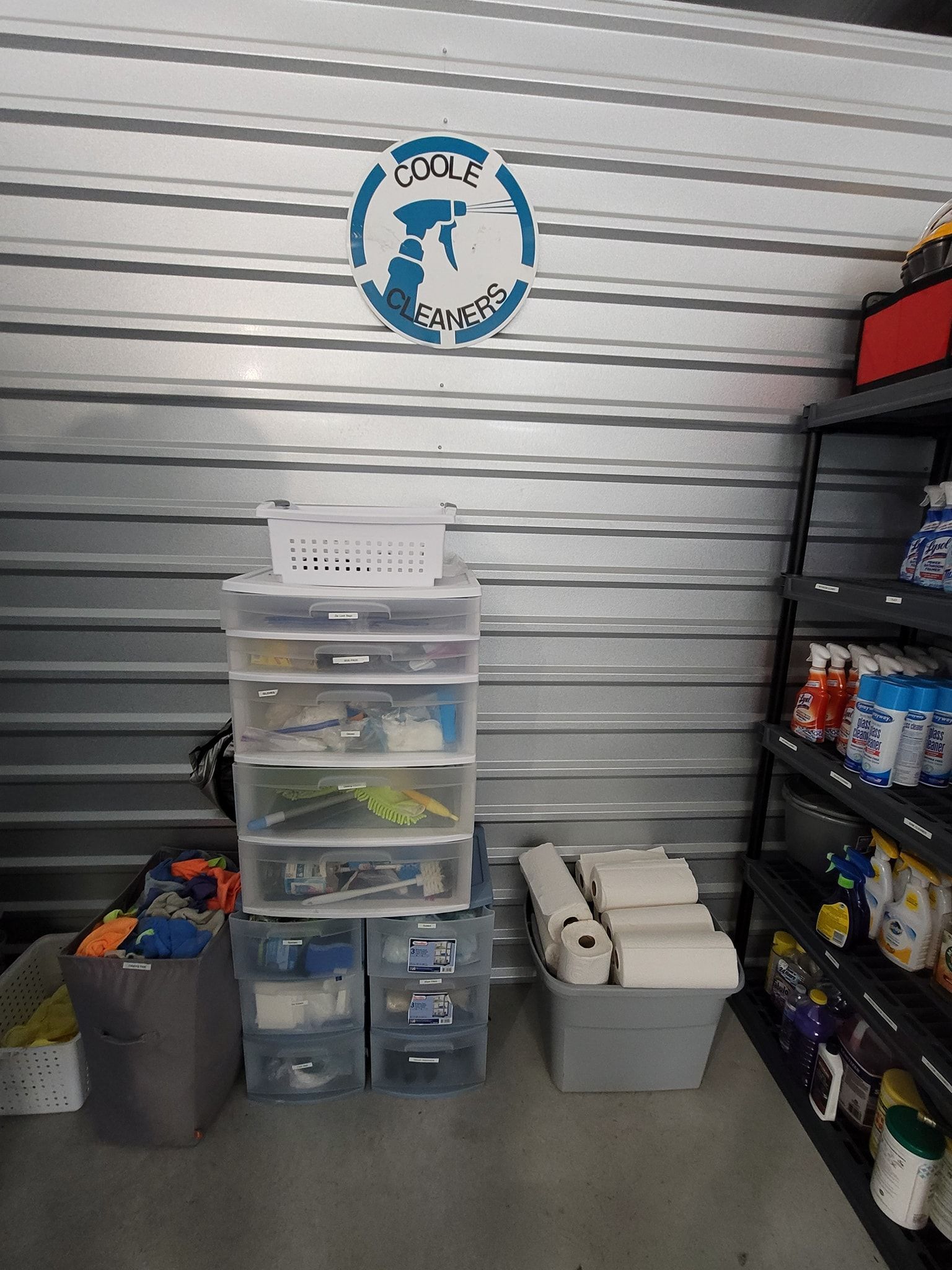 Storage unit with plastic drawers, shelves, and cleaning supplies. A sign is on the corrugated wall.